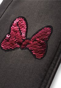 Écusson nœud rouge en sequins cousu sur un tissu texturé gris foncé avec des coutures visibles le long des bords.