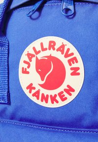 Modra tkanina nahrbtnik z ovalno belo oznako, ki vsebuje silhueto rdeče lisice in besedilo "FJÄLLRÄVEN KÅNKEN." Gladka tekstura, trdni zadrge.