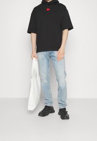 Schwarzer übergroßer Hoodie mit einem roten Etikett, kombiniert mit hellblauen Jeans. Hält eine weiße, strukturierte Tragetasche und trägt schwarze Sneakers.