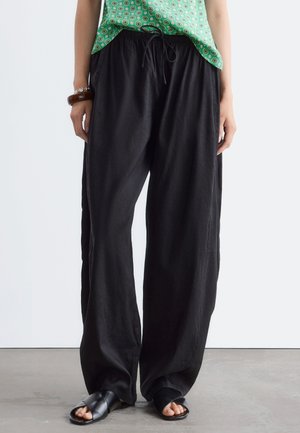 Person trägt weite schwarze Hose mit Kordelzug, ärmelloses grünes gemustertes Oberteil, schwarze Sandalen und ein braunes Armband und steht drinnen.