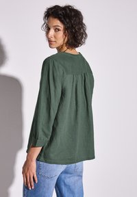 Top in lino verde con maniche lunghe e scollatura rotonda, caratterizzato da un dettaglio arricciato sul retro e una vestibilità rilassata. Abbinato a jeans azzurri chiari.