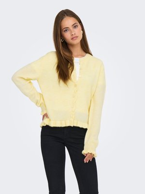 Joven con el pelo largo y castaño que lleva un cárdigan amarillo pálido abotonado y pantalones negros, de pie con una mano en la cadera.