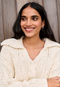 Elefántcsont színű, vastag kötésű pulóver gallérral és sodrott mintával. A modell hosszú sötét hajú, és mosolyog, miközben egy fa hátterű kép előtt áll.