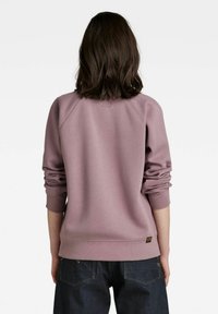 Osoba s hnědými vlasy po ramena, oblečená v mauve mikině s vyhrnutými rukávy a tmavě modrých džínách, otočená zády k fotoaparátu.