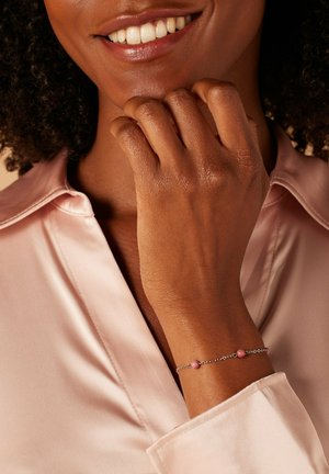 Zilveren kettingarmband met twee roze kralen, gedragen op een hand tegen een lichtroze satijnen blouse, die een gladde textuur en een delicate ontwerp toont.