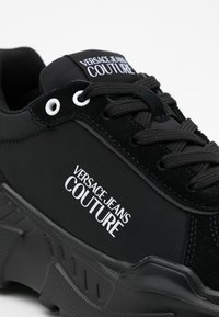 Baskets noires Versace Jeans Couture avec textures en tissu et daim, lacets noirs et texte du logo blanc sur le côté et la languette.