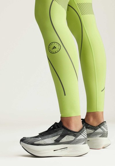 Neon green leggings with black accents, featuring a circular logo and curved lines. Black and grey running shoes with textured details.