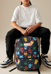 Svart ryggsäck med Marvel-superhjälte-design, med en framträdande logotyp, dragkedjor och färgglada karaktärstryck på en fläckig bakgrund.