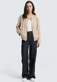 Beige bomberjack met elastische manchetten en taille, gedragen over een wit shirt en gecombineerd met brede donkeren denimbroeken en witte schoenen.