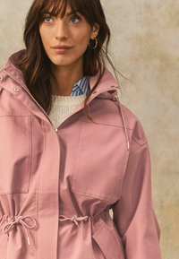 Helle pinke, wasserdichte Jacke mit Kordelzug in der Taille, einer Kapuze mit Druckknöpfen und zwei großen Fronttaschen. Strukturiertes Gewebe mit glatter Oberfläche.