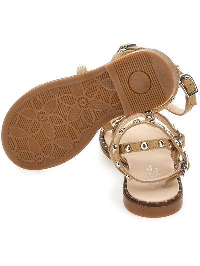 Sandalias de ante beige con suela plana, adornos de tachuelas plateadas y correa en el tobillo. La suela texturizada presenta un patrón floral.