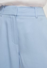 Pantalons bleu clair en tissu lisse, avec une taille plate, des poches latérales et des motifs de couture en diagonale.