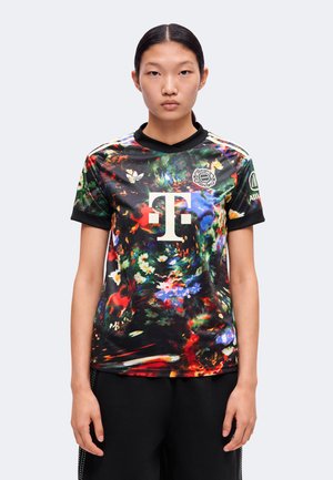 Personne portant un maillot de sport multicolore à motif abstrait avec un logo et un texte de sponsor, debout devant un fond clair uni.