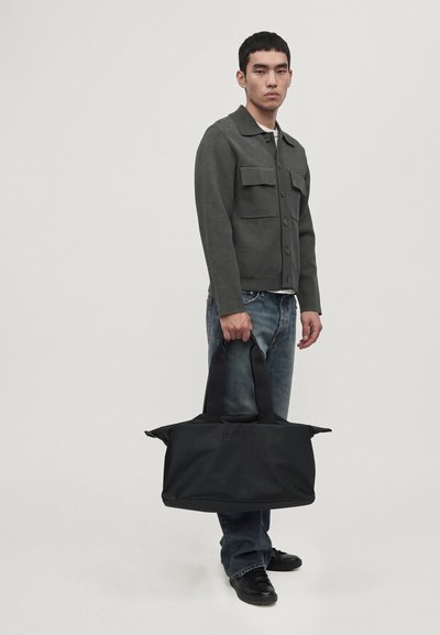 Un homme porte une veste boutonnée gris foncé et un jean, et tient un grand sac de voyage noir sur un fond clair uni.