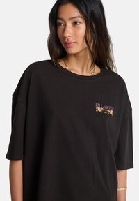 Μαύρο φαρδύ t-shirt από βαμβάκι. Διαθέτει μέτωπο με λογότυπο σε ροζ και κίτρινα λουλουδένια σχέδια. Κοντά μανίκια και στρογγυλή λαιμόκοψη.