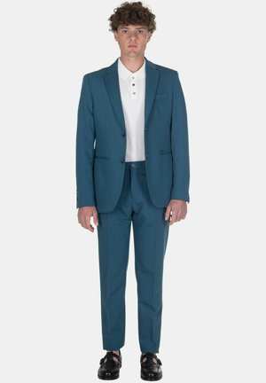 Giovane uomo con capelli ricci che indossa un completo turchese, una camicia bianca abbottonata e scarpe eleganti nere, in piedi davanti a uno sfondo semplice.