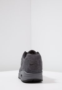 Sivé semišové tenisky s texturovaným povrchem, polstrovaným límcem a černými doplňky. Na zadní části je embosované logo Nike Air. Nízkoprofilový design.