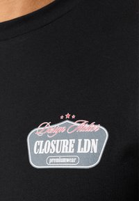 Svart bomullst-shirt med rundad halsringning och en framträdande dekorativ logotyp med text i vitt och rosa inom en grå sköldform.