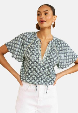 Kobieta ubrana w jasną wzorzystą bluzkę z falbankowymi rękawami i białe spodnie, pozująca z rękami na biodrach na jednolitym tle.