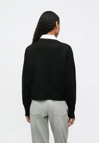 Maglione nero lavorato a maglia con design corto e maniche larghe, indossato sopra una camicia bianca con colletto, abbinato a jeans grigio chiaro.