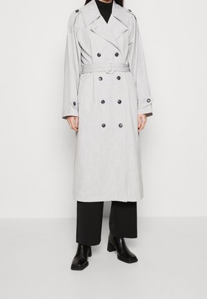 Femme portant un trench-coat gris clair croisé avec ceinture, pantalon noir à jambes larges et bottines noires à talons.