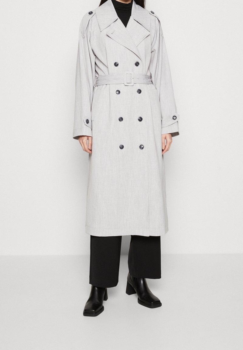 Femme portant un trench-coat gris clair croisé avec ceinture, pantalon noir à jambes larges et bottines noires à talons.