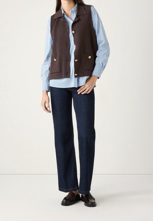 Frau trägt eine braune, geknöpfte Weste über einem blau-weiß gestreiften Hemd, dunkelblaue Jeans mit geradem Bein und dunkelbraune Loafer.