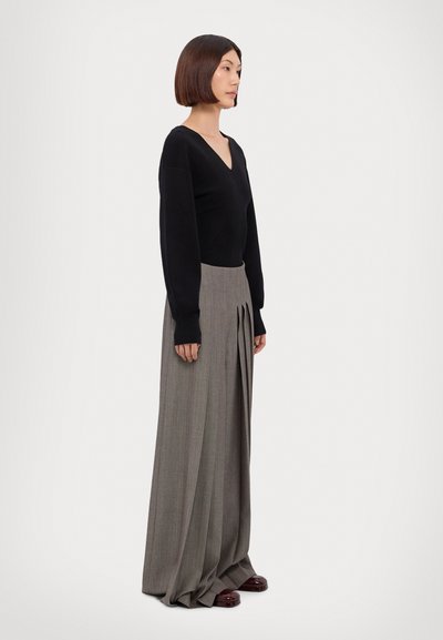 Haut noir à manches longues avec un col en V, associé à un pantalon beige plissé taille haute. Des chaussures bordeaux foncé complètent la tenue.