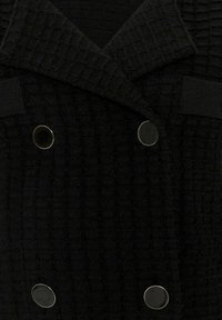 Manteau noir texturé à double boutonnage avec de larges revers et quatre gros boutons ronds noirs à bords argentés.