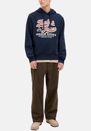 Uomo che indossa un hoodie blu navy con la scritta "Jack & Jones Denim Goods", pantaloni marroni, scarpe beige, in piedi davanti a uno sfondo bianco uniforme.