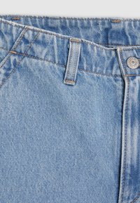 Primer plano de unos vaqueros de mezclilla azul claro que muestra la cinturilla, el ojal del cinturón, el botón metálico y una textura descolorida con costuras detalladas.