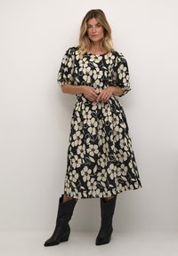 Musta mekko valkoisella kukkakuviolla, pyöreä pääntie, puffattu hihan, ja virtaava siluetti, yhdistettynä mustiin cowboy-kenkään.