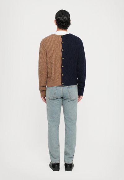 Stickad kofta med en delad design i brunt och marinblått, med ett kabelmönster, rundad halsringning och bakknäppning. I sällskap med ljusa jeans.