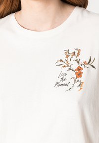 Camiseta de algodón blanca con un diseño floral que presenta flores naranjas y hojas verdes, impresa con "Vive el Momento" en texto negro.