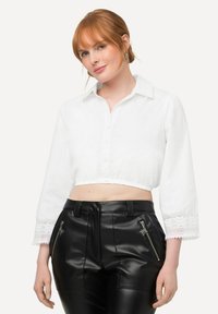 Weißes, kurz geschnittenes Blusenshirt mit langen Ärmeln, mit Spitzenbesatz an den Bündchen und einer Knopfleiste vorne. Kombiniert mit eng anliegenden schwarzen Lederhosen.