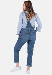 Denim overall, medium blauw, met verstelbare banden en opgerolde mouwen. Gecombineerd met een lichtblauw gestreept overhemd met knopen en witte sneakers.