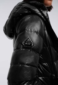 Primo piano di una spalla su un giubbotto di piumino nero lucido con cappuccio, che mostra una toppa del marchio triangolare JK1T.