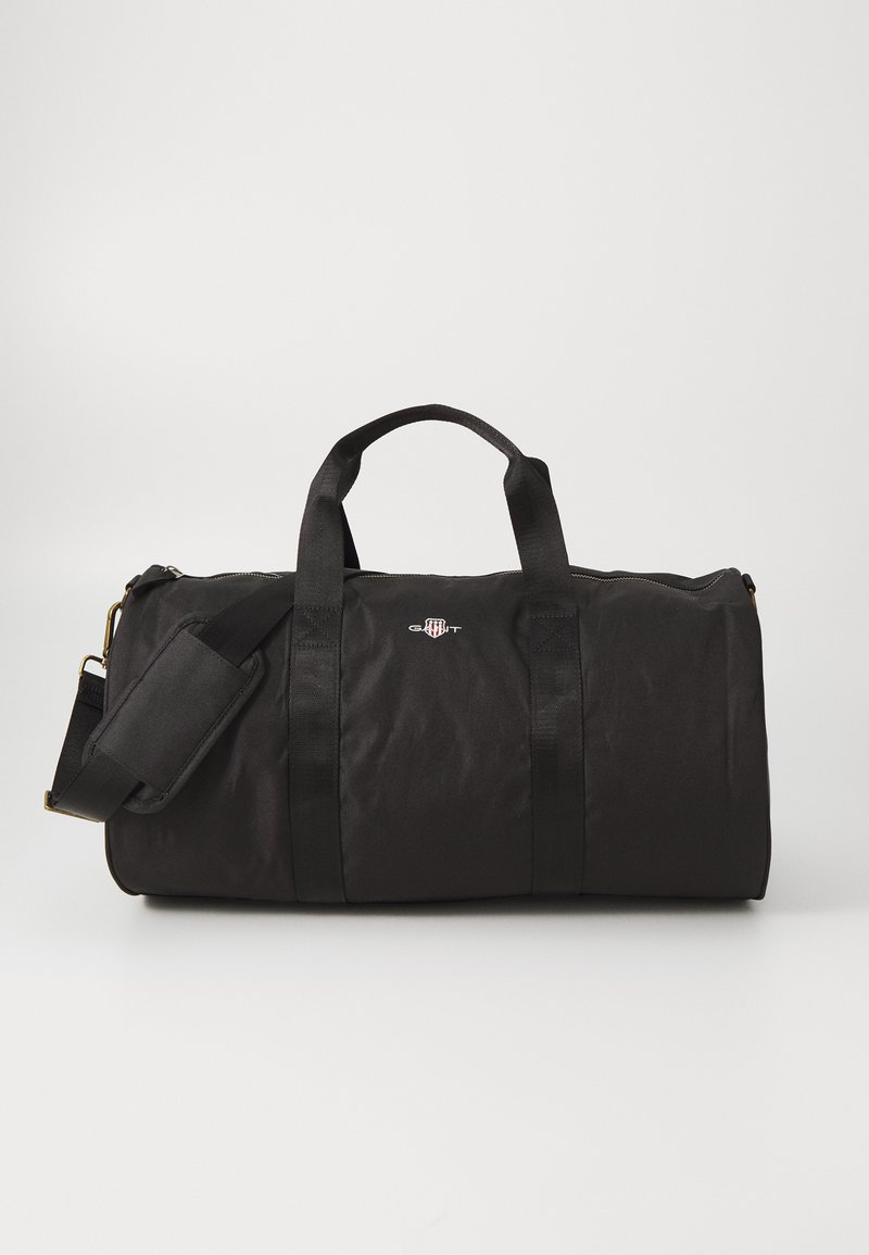 Svart duffelbag laget av slitesterkt stoff, med to topphåndtak, en justerbar skulderrem, og en frontlogo.
