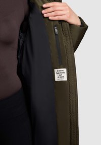 Olijfgroene pufferjack met een zwarte voering, voorzien van een ritszak en een label met de tekst "Een warme omhelzing gemaakt van stof."
