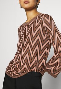 Pitkähihaisen neuleen, jossa on vaaleanpunaisia ja ruskeita sävyjä käyttäjä tikkukuvio. Pehmeä kangas, jossa on hieman rypytetty helma ja ribbiret. 