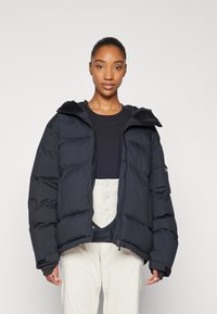 Puffer jacket in black, featuring a zip closure, wide padded sections, and a detachable hood. Worn with a dark top and light-colored bottoms.