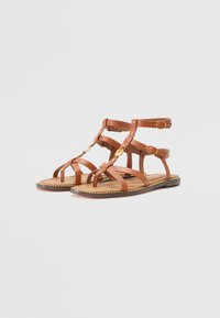 Brune lær sandaler med flere stropper og flad sål. Har en vævet kant og hardware i guldfarve.