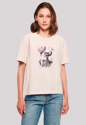 Hellrosa Baumwoll-T-Shirt mit einem Skelett, das einen herzförmigen Ballon hält, umgeben von dekorativen Mustern und Schmetterlingen.