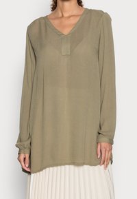 Blouse à manches longues vert olive avec un col en V, un détail plissé à l'avant, et une coupe ample ; fabriquée dans un tissu léger et texturé.