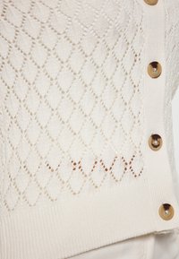 Cardigan tricotat de culoare crem cu un model de decupaj în formă de diamant, tiv cu nervuri și nasturi mari, rotunzi, cu un finisaj lucios.