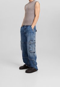 Grå ribbstickad ärmlös topp i kombination med lösa blå denim cargobyxor med sidofickor och en urtvättad look. Svarta slip-on skor.