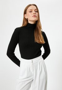 Svart ribbad polotröja med långa ärmar och en figurnära design. Matchas med vita byxor med hög midja, som visar en slät tygkvalitet och skräddarsydd look.