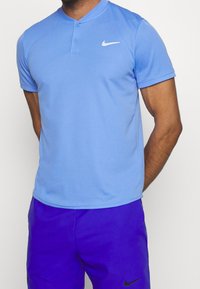 Camisa polo de Nike de color azul claro con mangas cortas y cuello tipo henley, con un logo blanco y un tejido suave y transpirable. Combinada con pantalones cortos morados.