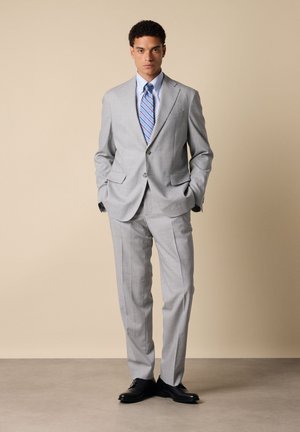 Man staat met handen in de zakken, draagt een lichtgrijs pak, een blauw gestreepte stropdas, een lichtblauw overhemd en zwarte nette schoenen tegen een beige achtergrond.