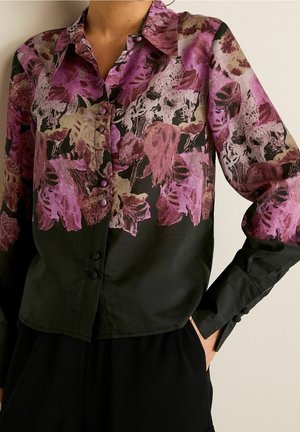 Blouse boutonnée à manches longues avec imprimé floral violet et rose sur la moitié supérieure et uni noir sur la moitié inférieure, portée avec un pantalon noir.